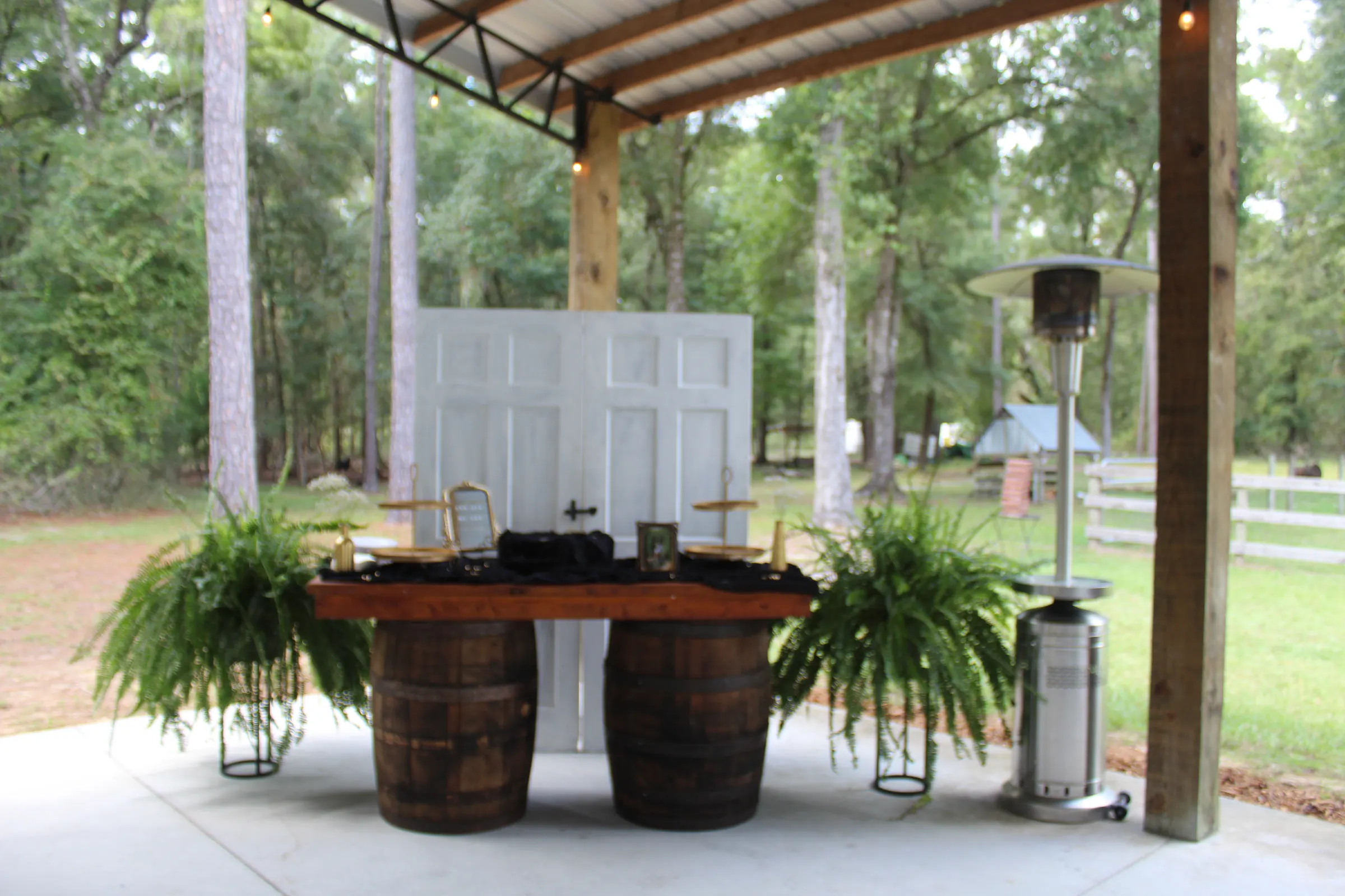 A serving bar made from a plank top laid across two whiskey barrels, hanging ferns on either side, standing on the pavilion's concrete floor with white doors behind it and trees past the open edge.