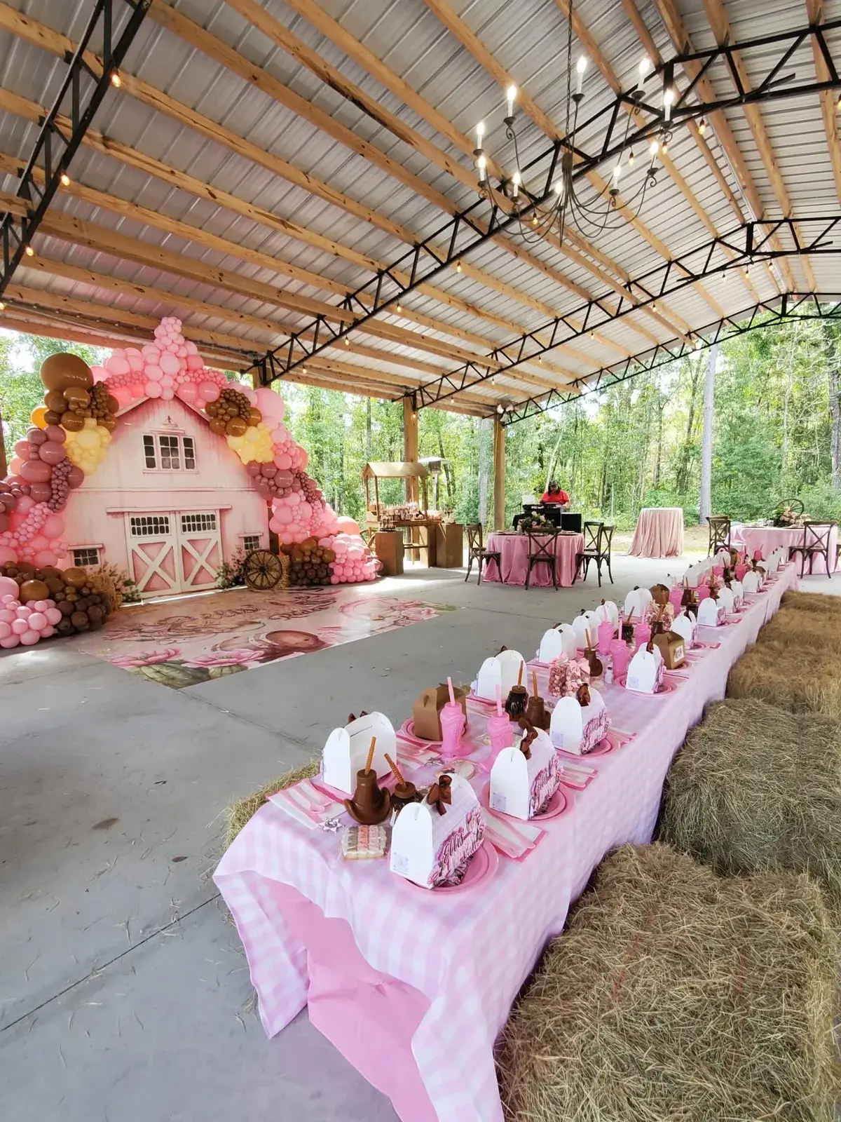 A long table under a pink gingham cloth running down the pavilion floor, set with place settings and lined with straw bales, a pink barn backdrop and balloon garland at the head of it.