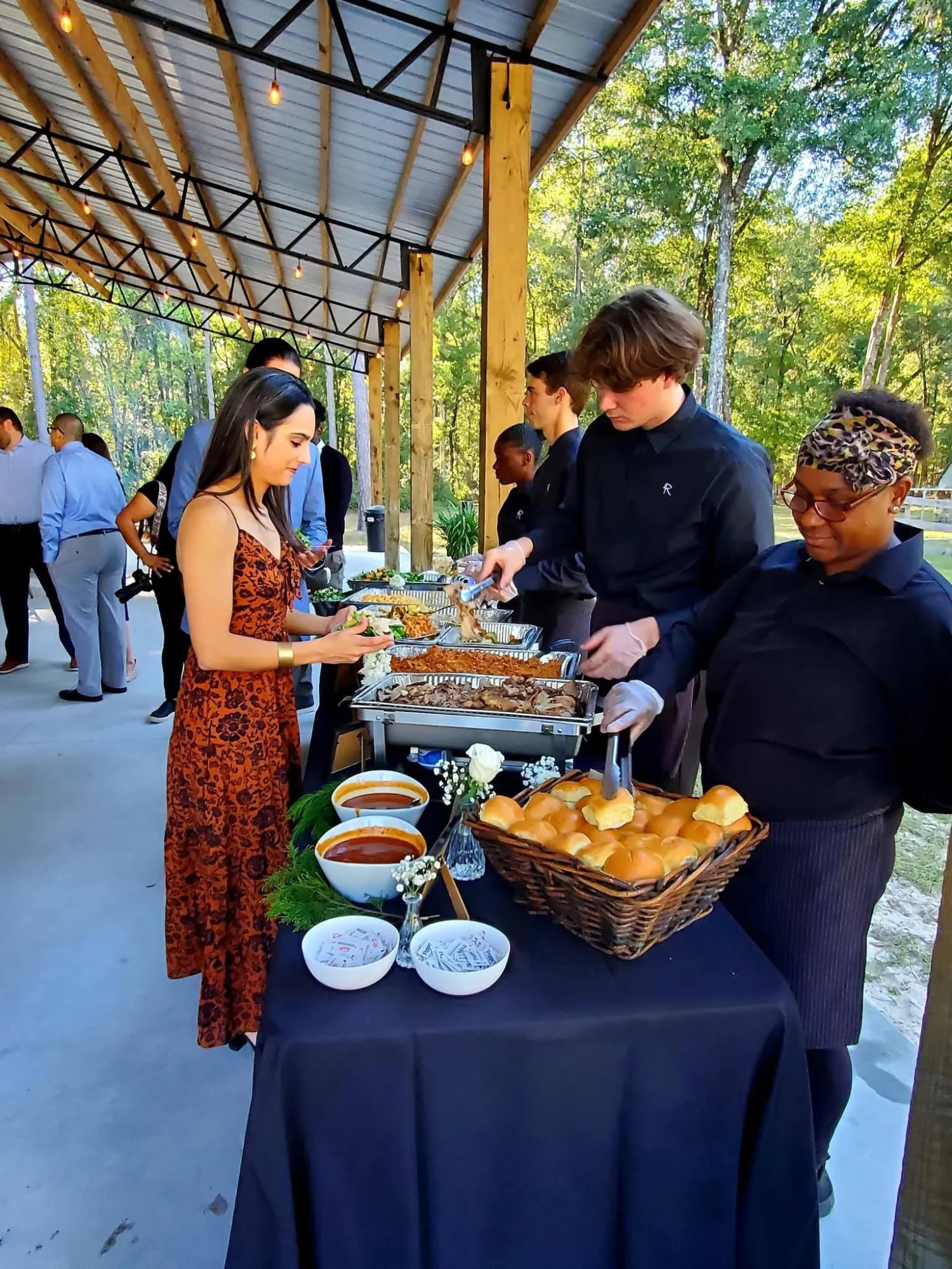 Two catering staff in black serving from chafing dishes on a long black draped buffet under the open pavilion, a guest in an orange floral dress filling her plate, a basket of rolls in the foreground and more guests talking in the background.