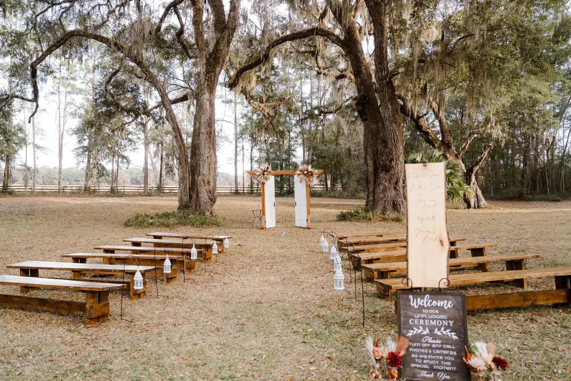 Rows of long wooden benches set on open grass facing a timber arbor, white lanterns hanging from shepherd hooks along the aisle and a chalkboard welcome sign in the foreground.
