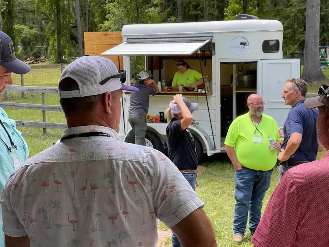 A group of men in caps standing on the grass around the white horse trailer bar, a server leaning out through its serving window, pasture fencing and pines behind.