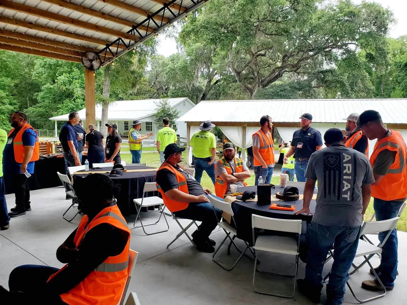 Men and women in orange and yellow high-visibility work vests sitting and standing around folding tables under the open pavilion, with white farm buildings and oaks behind them.