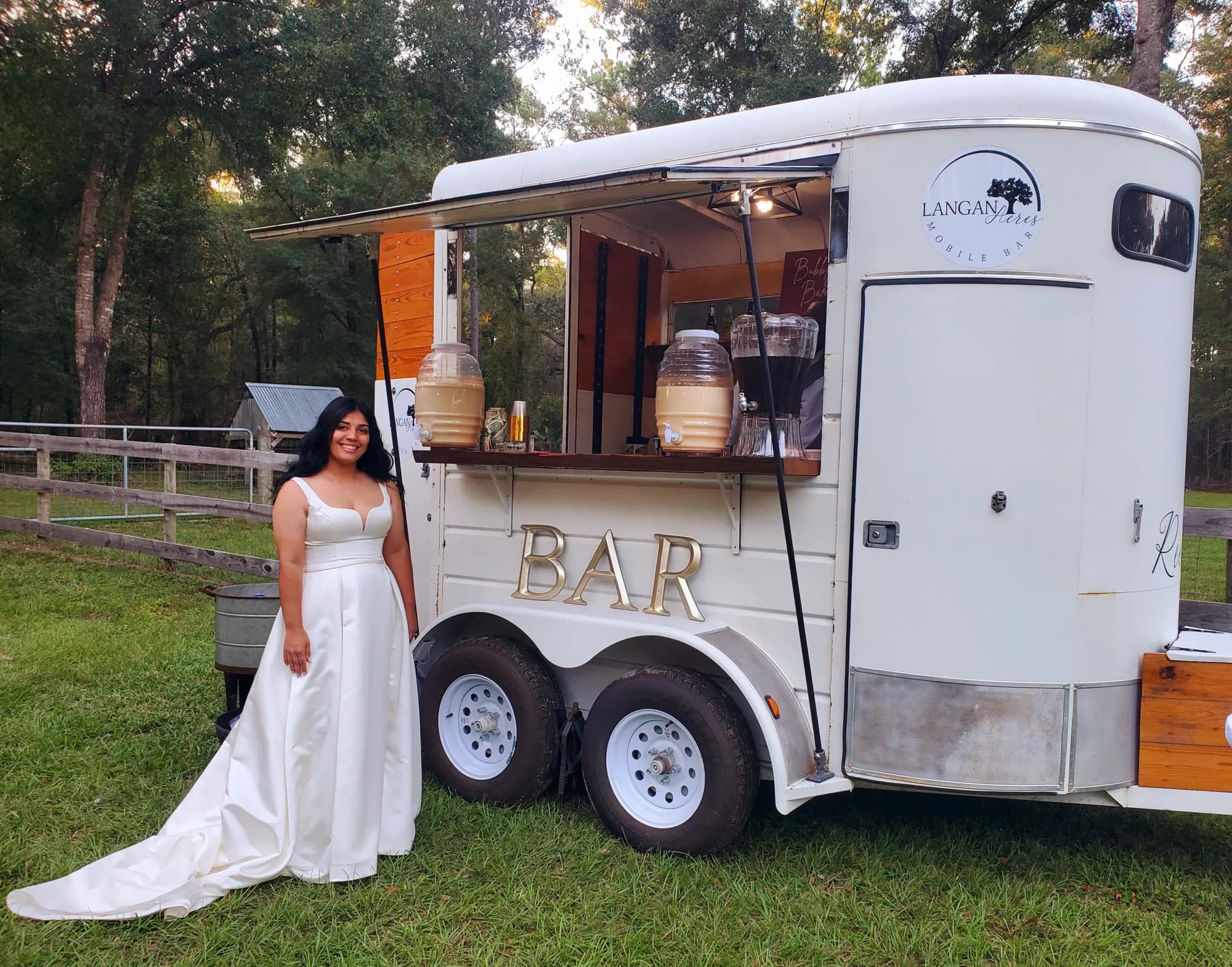 A bride in a white satin gown standing on the grass beside the white mobile bar trailer, its hatch propped open and lit, gold BAR lettering on the side, two glass drink dispensers on the counter and the Langan Acres Mobile Bar logo on the door.