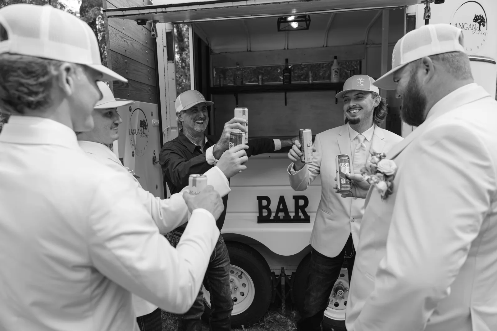 Black and white photograph of a group of men in pale suits and ball caps standing in a circle in front of the open mobile bar trailer, raising cans together in a toast, with the Langan Acres Mobile Bar logo and the word BAR on the trailer behind them.