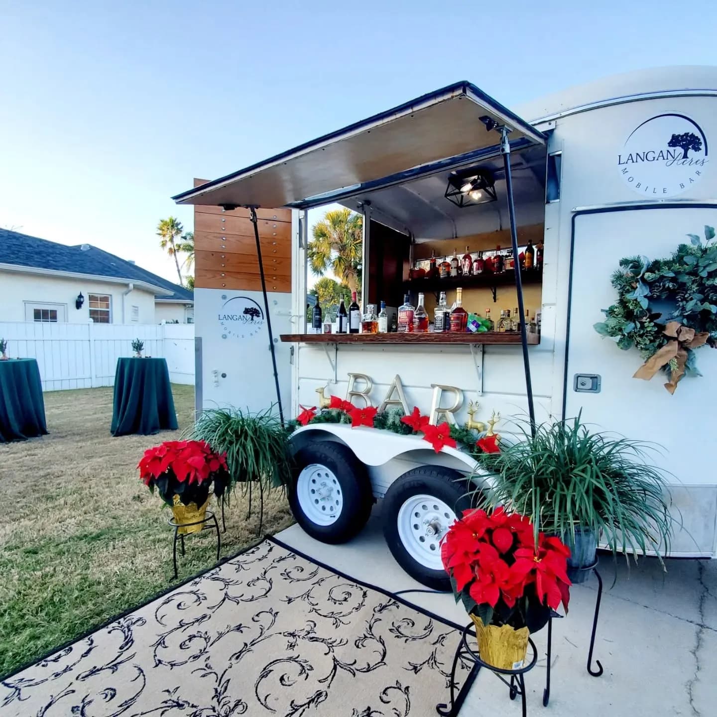 The white mobile bar trailer set up on a driveway at a private house, its hatch open over a counter of spirit bottles, dressed for Christmas with a greenery wreath on the door, red poinsettias in gold pots and a garland along the gold BAR lettering, with cocktail tables in green cloths on the lawn behind.