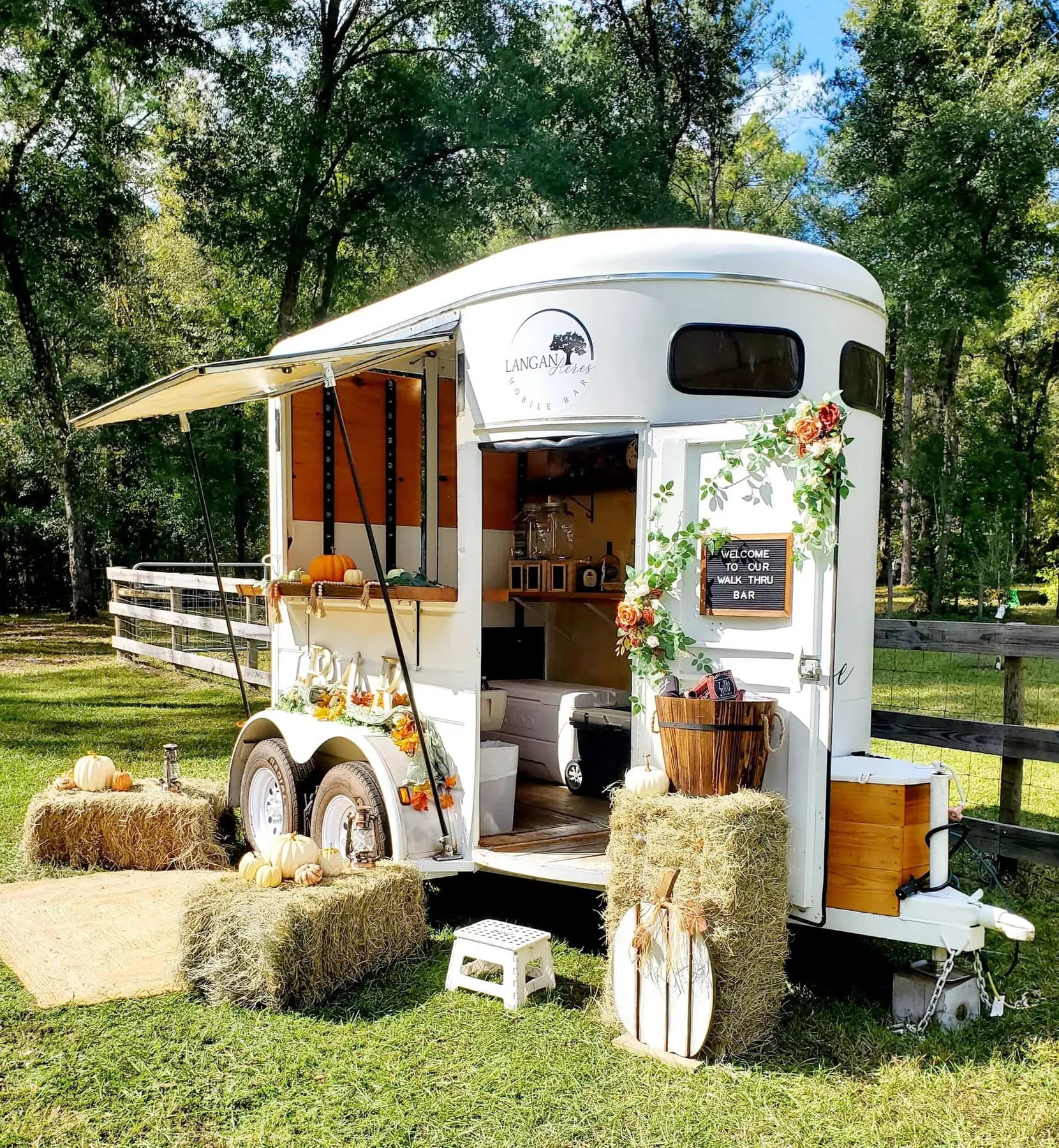 A white horse trailer converted into a bar, awning extended, serving window open onto timber shelving, dressed with pumpkins, hay bales, lanterns and an autumn garland, a letterboard beside the door reading welcome to our walk thru bar.