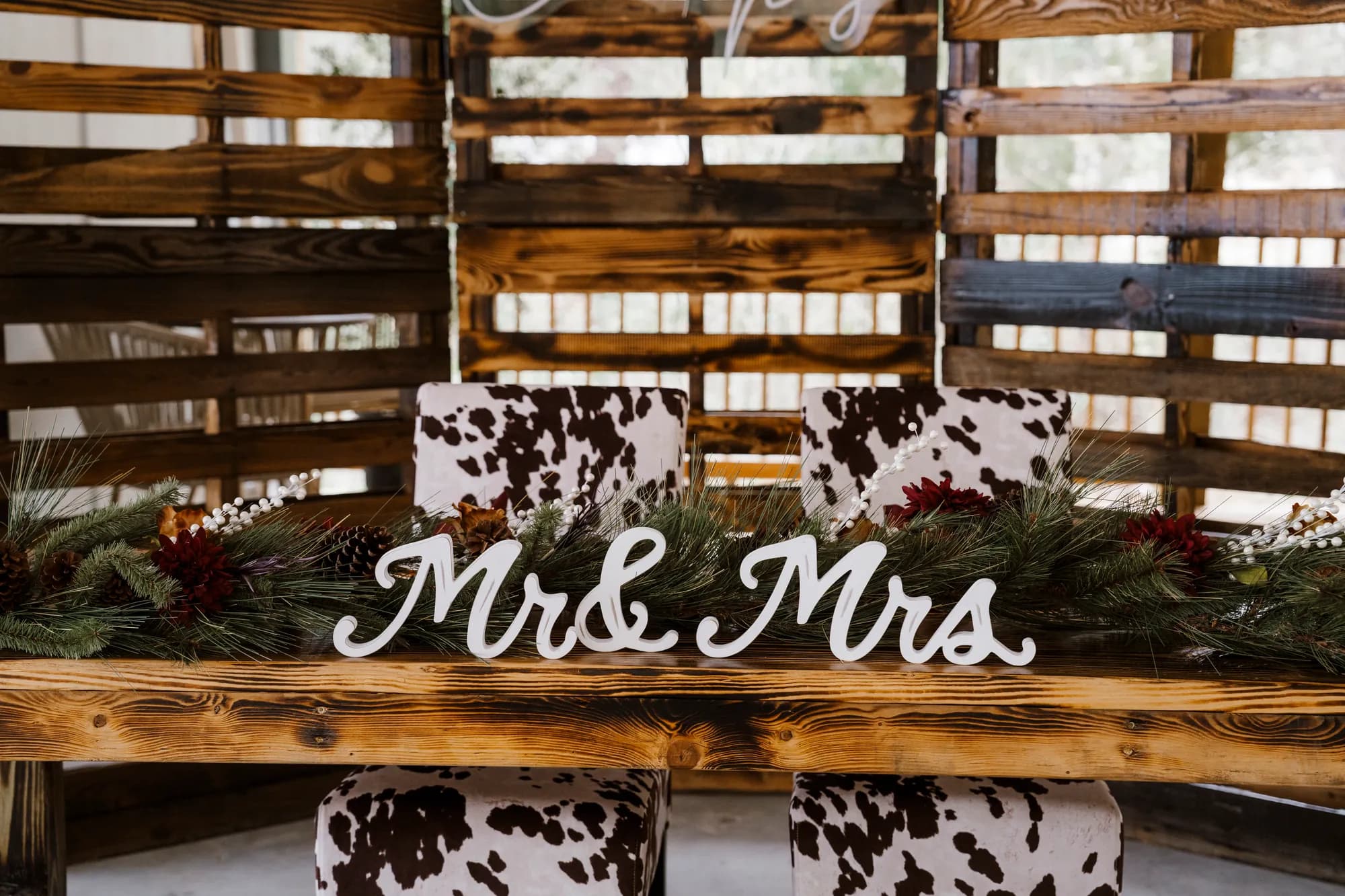 A rustic wood sweetheart table with a Mr and Mrs sign, cowhide print chairs and pine and berry garland, set against a stained pallet wall.