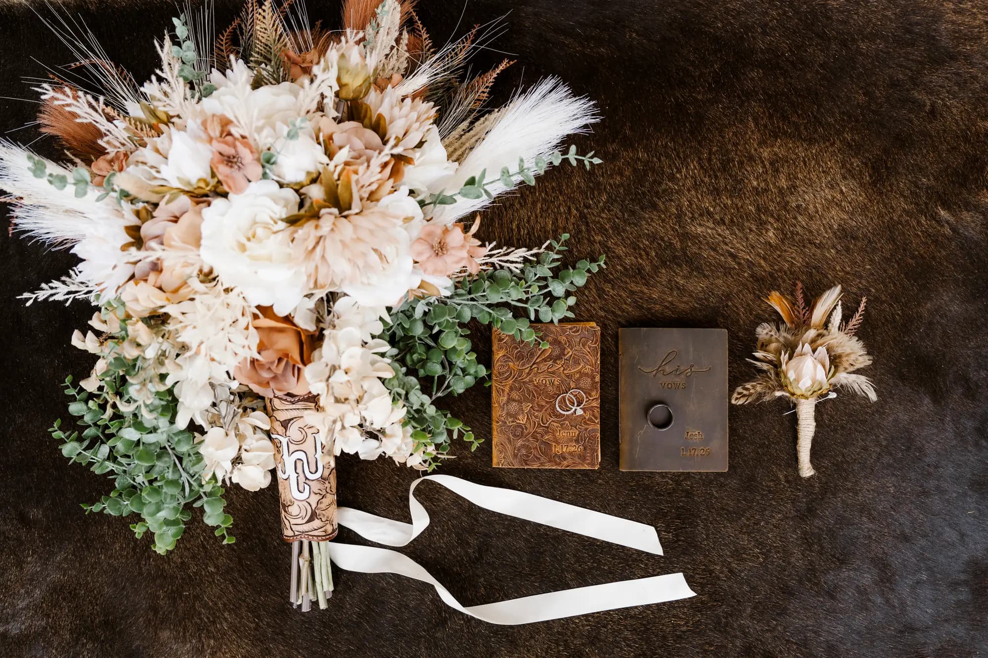 A bouquet of cream and rust coloured dried flowers, two tooled leather vow books and a matching boutonniere arranged on a brown cowhide.