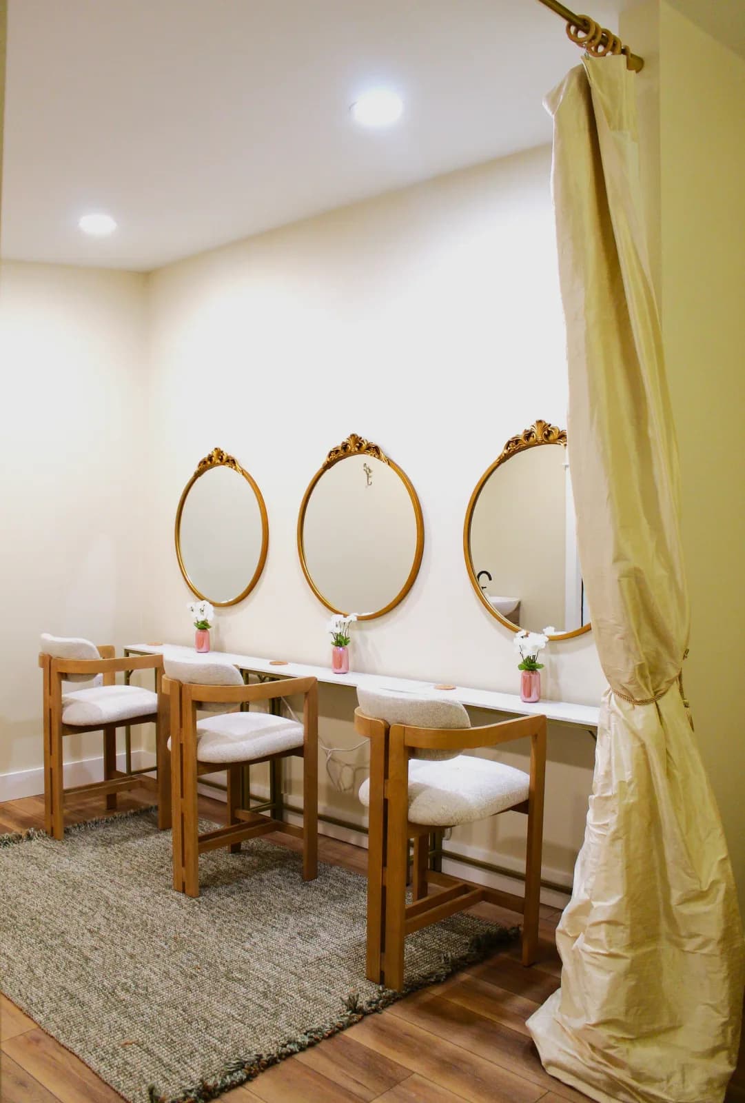 Three gold oval mirrors above a long white getting-ready counter, with upholstered stools and small vases of flowers