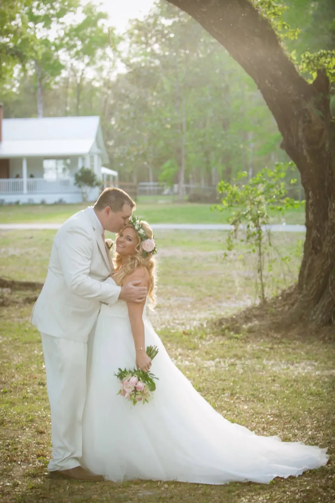 A bride and groom in white, holding hands as they walk beneath the oaks