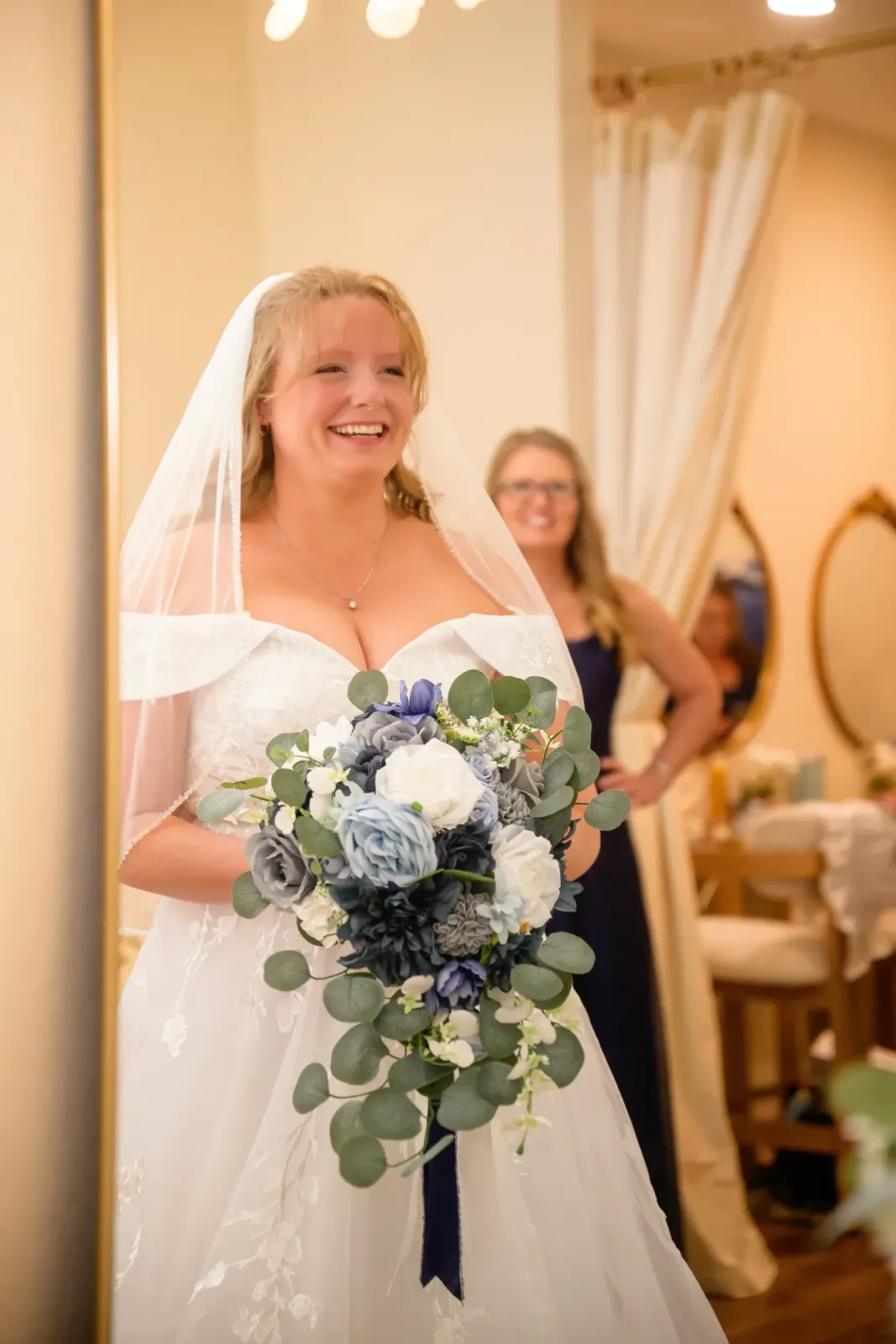A bride in a veil holding a blue and white bouquet in the bridal suite, a bridesmaid and the room's mirror behind her.