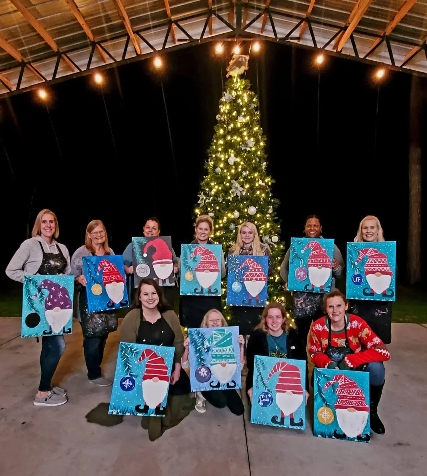 A painting class holding up their finished canvases together in front of a lit Christmas tree
