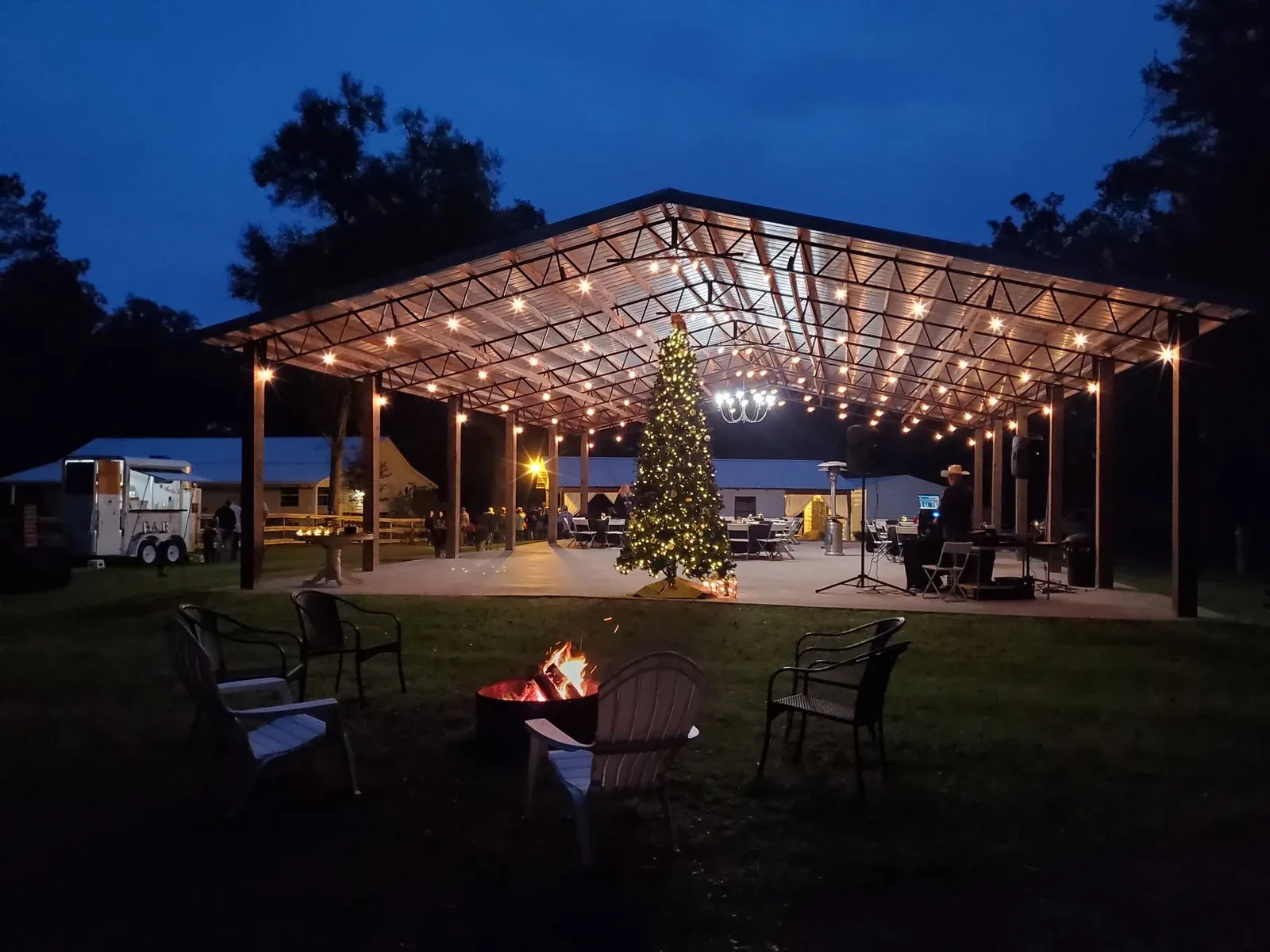 The open-air pavilion strung with warm lights at dusk, a lit Christmas tree inside and a fire pit burning in the foreground