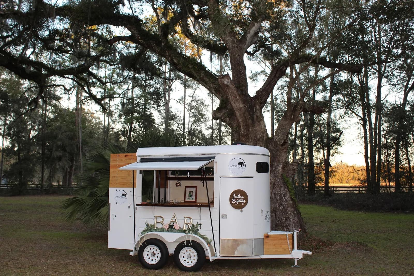 Reese, the Langan Acres mobile bar: a white horse trailer converted to a bar, serving hatch open with wood shelving and a BAR sign, parked under a live oak at dusk.