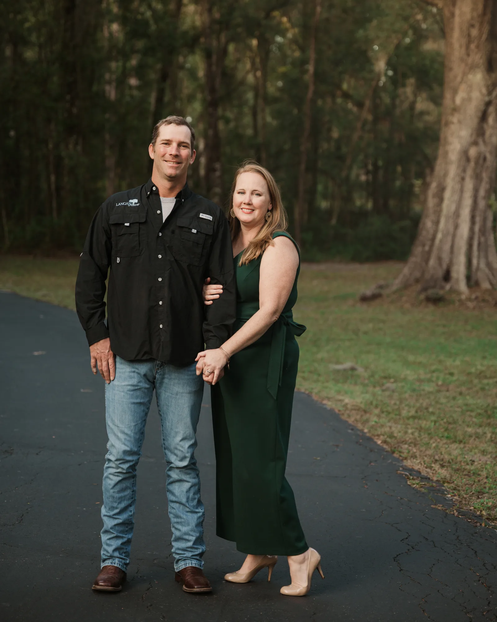 Adam and Betsy Langan standing hand in hand on the paved drive at Langan Acres, Adam in a dark work shirt and jeans, Betsy in a long forest green dress, live oaks and pines behind them.