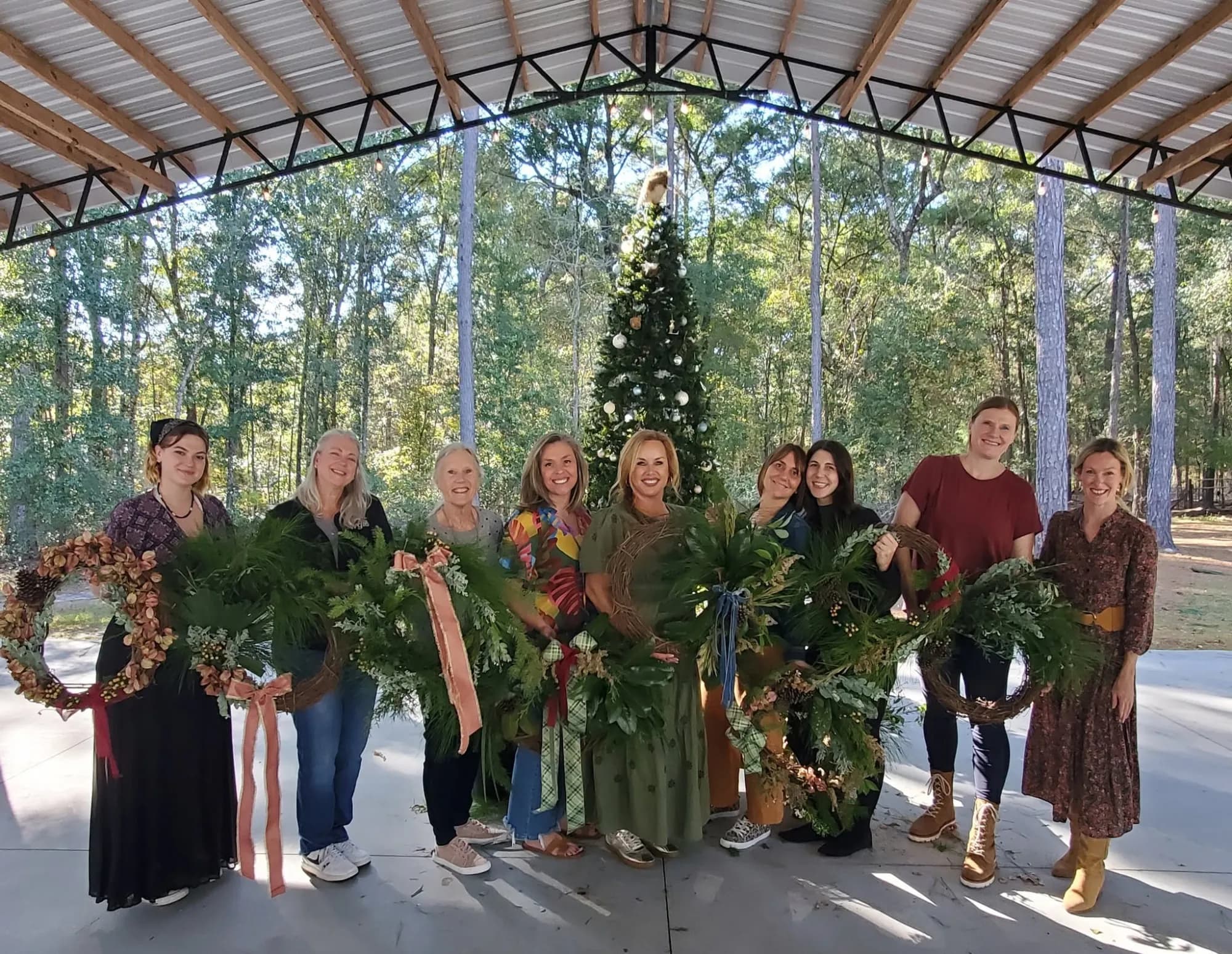 Nine women standing shoulder to shoulder under the open pavilion, each holding up the greenery wreath she made, a decorated tree and the surrounding woods behind them.