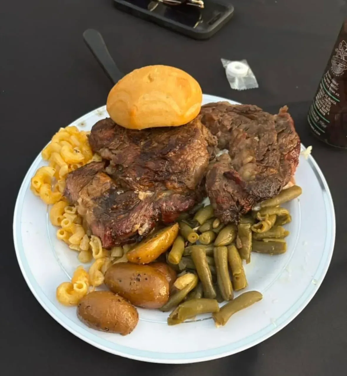 A paper plate of grilled steak with macaroni cheese, green beans, roast potatoes and a bread roll, on a dark table at the Steers & Beers fundraiser.